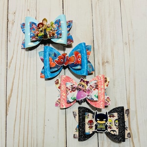 May include: Four handmade hair bows with different cartoon character designs. The bows are made of fabric and have a glitter finish. The characters include Toy Story, Finding Nemo, Elmo, and Batman.