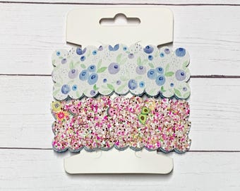 May include: Two packs of scalloped edge fabric trim. The top pack features a white background with a blue blueberry pattern. The bottom pack features a pink glitter background with a watermelon, avocado, and lemon pattern.