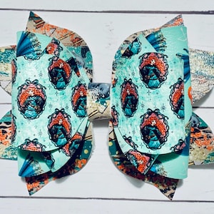 May include: A teal blue hair bow with a layered design featuring a repeating pattern of a cartoon princess with red hair. The bow has a glittery finish.