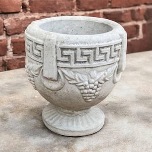 May include: A gray concrete planter with a Greek key design and grapevine motif. The planter has a wide base and a narrow opening.