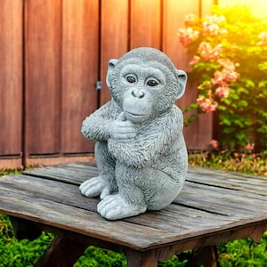 May include: A gray monkey statue sitting on a wooden table. The statue is in a seated position with its hands clasped. The background includes a wooden fence and greenery.
