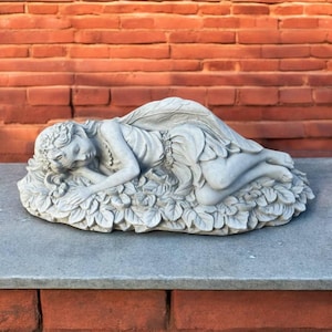 May include: A gray stone fairy statue in a sleeping pose, resting on a bed of leaves and flowers. The fairy has detailed wings and a floral crown. The statue is displayed against a red brick wall.