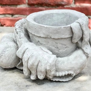 May include: A gray concrete planter shaped like a baseball player holding a baseball and a glove. The planter has a round opening at the top for planting.