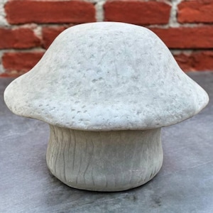 May include: A light gray mushroom-shaped garden ornament. The mushroom has a rounded cap and a textured surface. The stem is cylindrical with vertical grooves. The background features a brick wall.