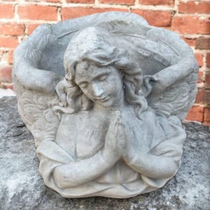 May include: A gray angel statue with folded hands and large wings. The angel is in a praying position, with a serene expression. The statue is made of a stone-like material and is likely a garden ornament or decorative piece.
