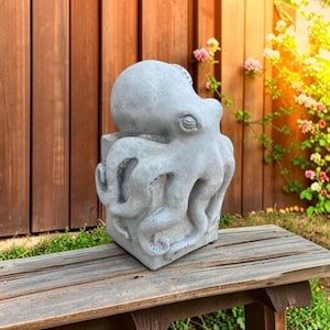 May include: A light gray concrete sculpture of an octopus with a rounded head and multiple textured tentacles wrapped around a square base. The sculpture features a weathered finish with visible details on the eyes and suckers.