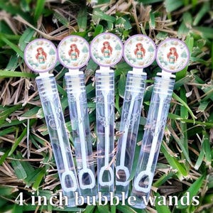 May include: Five clear plastic bubble wands with white caps, each featuring a circular topper with a printed illustration of a red-haired mermaid character. The wands are cylindrical and transparent, showing the internal bubble blowing mechanism.