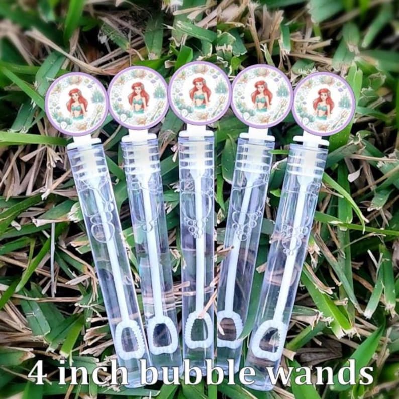 May include: Five clear plastic bubble wands with white caps, each featuring a circular topper with a printed illustration of a red-haired mermaid character. The wands are cylindrical and transparent, showing the internal bubble blowing mechanism.