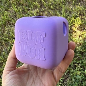 May include: A light purple, cube-shaped container with rounded edges. The front face features the embossed text "ELF DOH" in a stylized, wavy font. The object has a circular opening on the top and a circular hole on the right side panel.