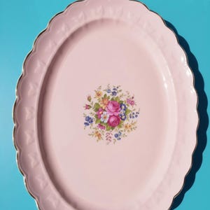 May include: An oval-shaped light pink serving platter with a scalloped edge and a gold-toned rim. The centre of the platter features a detailed floral bouquet illustration containing pink, yellow, blue, and green flowers and foliage.