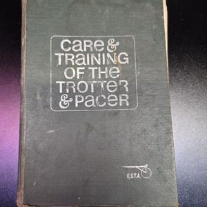 May include: A dark green hardcover book titled Care & Training of the Trotter & Pacer with white text on the front cover. The cover features a small white graphic of a sulky and the letters U.S.T.A. in the bottom right corner of the book.