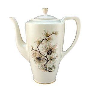 May include: A white ceramic coffee pot with a gold trim and a pine tree design. The pot has a lid and a handle.