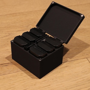 May include: A black box with a hinged lid, open to reveal six black, curved compartments. The box is on a wooden surface. The lid is open, revealing the interior. The box appears to be made of a matte material.