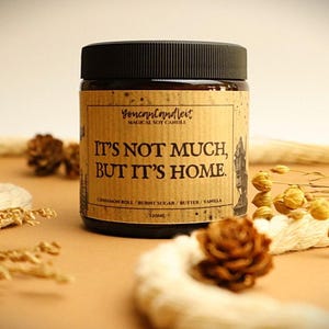 Op de afbeelding: Een sojakaars in een glazen pot met een zwart deksel. Het etiket luidt "IT'S NOT MUCH, BUT IT'S HOME." met geurnoten van kaneelbroodje, gebrande suiker, boter en vanille. De pot is 120 ml.