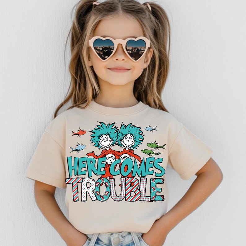 May include: A light beige t-shirt with a graphic of the Thing 1 and Thing 2 characters from Dr. Seuss's book, "The Cat in the Hat". The graphic includes the text "Here Comes Trouble" in red, white, and blue. The shirt has short sleeves and a crew neck.