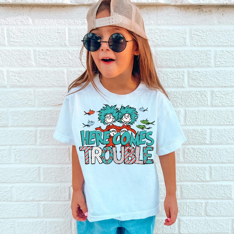 May include: A white t-shirt with a graphic of two green-haired figures with red shirts and the text "HERE COMES TROUBLE" in red and white stripes. The figures are surrounded by blue and green fish.