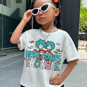 May include: White short-sleeved t-shirt with a graphic print of two characters from the book "The Cat in the Hat" and the text "Here Comes Trouble".