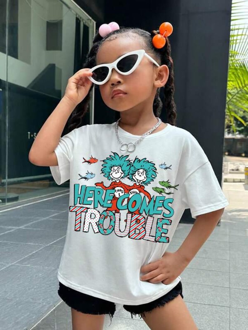 May include: White short-sleeved t-shirt with a graphic print of two characters from the book "The Cat in the Hat" and the text "Here Comes Trouble".