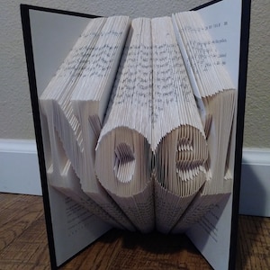 Peut inclure: Une sculpture d'art de livre du mot "HOME" créée en pliant les pages d'un livre. Le livre est ouvert et les pages sont pliées pour créer les lettres. Le livre est posé sur une surface en bois.