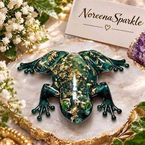 May include: A decorative frog figurine in a deep emerald green colour with embedded gold leaf flakes and bright green eyes. The frog is positioned on a white stone slab with a gold-trimmed edge, accompanied by a card reading Noreena Sparkle.