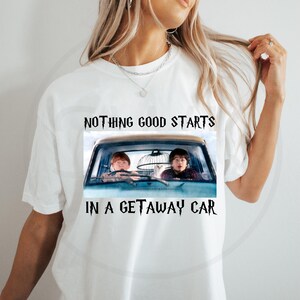 Op de afbeelding: Wit T-shirt met een afbeelding van twee personages in een blauwe auto en de tekst "NOTHING GOOD STARTS IN A GETAWAY CAR" in het zwart. De afbeelding is een filmreferentie.