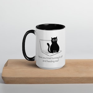 Könnte beinhalten: Eine weiße Kaffeetasse mit schwarzem Rand und Henkel. Die Tasse zeigt eine schwarz-weiße Illustration einer Katze, die auf einem Laptop sitzt, mit dem Text "Hast du versucht, ihn auszuschalten und mich zu füttern?"