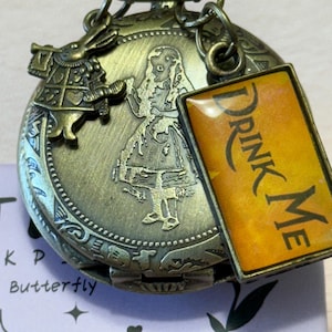 May include: Vintage bronze pocket watch with an engraved Alice in Wonderland design, including the White Rabbit and Alice. A rectangular charm with the words "DRINK ME" is attached. The watch has intricate detailing and a classic, antique appearance.