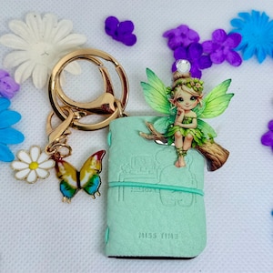 May include: A mint green journal with a fairy figurine, butterfly and daisy charms, and a gold keychain. The fairy has green wings and is perched on a branch. The journal has the text "MISS TIME". Decorative flowers surround the items.