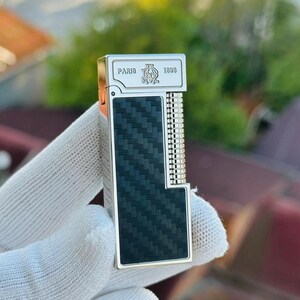 May include: A silver and black lighter with a carbon fibre pattern. The top of the lighter has the text "PARIS 1898" and a decorative emblem. The lighter is held in a white glove.