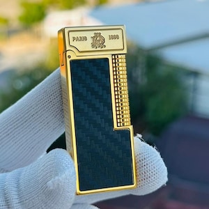 May include: A gold-coloured lighter with a black textured panel and a grid-like design on the side. The lighter has the word "PARIS" and the year "1898" engraved on the top.