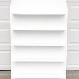 May include: A tall, white, freestanding display shelf featuring a smooth, arched top design. The unit includes four horizontal shelves spaced evenly, providing multiple levels for storage or presentation. The structure is finished in a solid matte white.