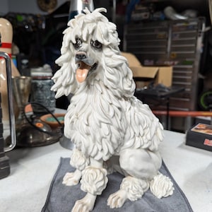 Op de afbeelding: Een wit porseleinen beeldje van een zittende poedel op een grijze doek. De poedel heeft een pluche witte vacht en een zwarte neus. De tong van de hond steekt uit.