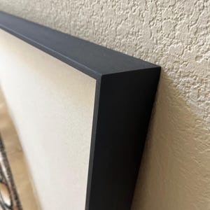 May include: Close-up of the corner of a black picture frame with a beige mat and a white background.