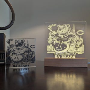 May include: Two illuminated acrylic signs featuring a bear wearing a football jersey with the number 1 and the word "BEARS". The bear is smoking a cigar and holding a beer mug. The signs are on a wooden base and have the words "DA BEARS".