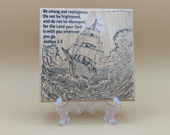 May include: Wooden plaque with a black line drawing of a sailboat navigating rough seas. The plaque features the text "Be strong and courageous...Joshua 1:9". The artwork is displayed on a clear plastic easel.