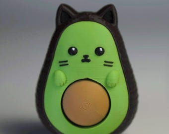May include: A small figurine shaped like an avocado with cat ears and a face. The outer shell is dark brown, the inner flesh is bright green, and the central pit is a light tan colour. The cat features include pointed ears, whiskers, and a nose.