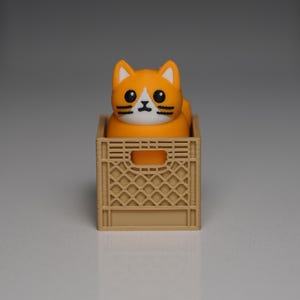 Puede incluir: Una figura de gato naranja y blanco con detalles negros se encuentra dentro de una peque&ntilde;a caja de color beige. El gato tiene grandes ojos negros y una mancha blanca en la cara. La caja tiene un dise&ntilde;o de celos&iacute;a y una abertura rectangular en el lateral.