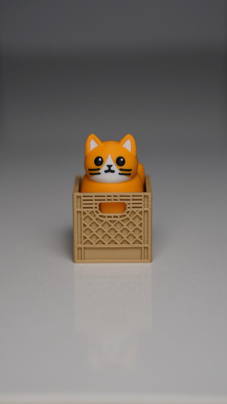 Puede incluir: Una figura de gato naranja y blanco con detalles negros se encuentra dentro de una peque&ntilde;a caja de color beige. El gato tiene grandes ojos negros y una mancha blanca en la cara. La caja tiene un dise&ntilde;o de celos&iacute;a y una abertura rectangular en el lateral.
