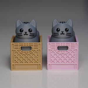 Puede incluir: Dos figuras de gato gris sentadas dentro de cajas en miniatura, una beige y otra rosa. Los gatos tienen ojos y bigotes negros. Las cajas tienen un diseño de celosía y una abertura rectangular en la parte delantera.