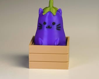 May include: A purple aubergine-shaped cat figurine with a green stem on top, black eyes, whiskers, and a small mouth. The figurine is seated inside a small, light brown, rectangular crate with horizontal slats on the sides and an open top.