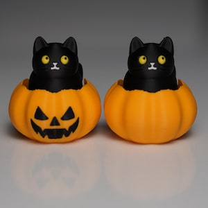 May include: Two black cat figurines with yellow eyes sit inside orange jack-o'-lanterns. The pumpkins have carved faces with triangular eyes and a toothy grin. A Halloween-themed decoration.