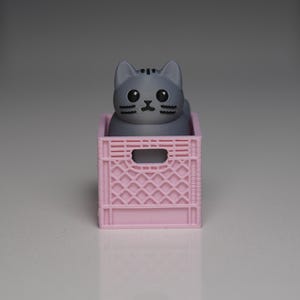 May include: A grey cat figure with big eyes is in a small pink plastic crate. The cat has a detailed face and is in the centre of the crate. The crate has a rectangular opening on the front and is made of a lattice-like design.