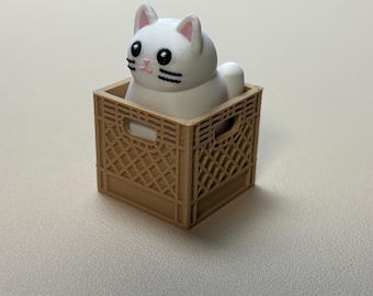 May include: A small white cat figurine with black eyes, black whiskers, and pink inner ears, sitting inside a tan-coloured plastic crate with a grid-like pattern on its sides. The cat is positioned upright, filling the interior of the crate.