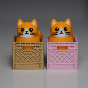 Puede incluir: Dos figuras de gato naranja con caras blancas y ojos negros sentadas en cajas en miniatura. Una caja es de color canela, la otra es rosa. Los gatos tienen bigotes negros y son de dise&ntilde;o lindo y caricaturesco.