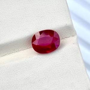 Può includere: Una gemma dal taglio ovale di un colore rosso profondo e saturo, con molteplici sfaccettature triangolari che creano una superficie brillante e riflettente. La pietra presenta un'elevata purezza e una finitura lucida, mostrando una ricca tonalità cremisi.