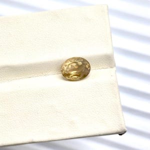 Puede incluir: Una piedra preciosa facetada de forma ovalada con un tono amarillo dorado translúcido. La piedra presenta un corte brillante con múltiples facetas triangulares y en forma de cometa que reflejan la luz, creando un aspecto brillante.