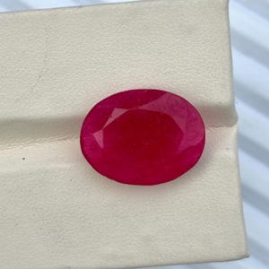 May include: A vibrant, oval-cut ruby gemstone with a deep red hue and multifaceted surface. The stone features a polished finish that reflects light across its top, showcasing a clear, saturated color profile and a symmetrical, rounded oval shape.