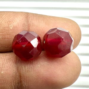 May include: Two round, faceted beads in a deep, translucent red color. Each bead features multiple flat, geometric surfaces that catch the light, creating a sparkling effect. The beads are small, spherical, and have a polished, glass-like finish.