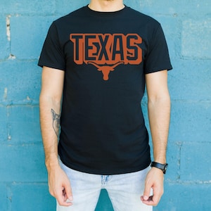 May include: Black t-shirt with the word "TEXAS" in burnt orange block letters and a longhorn logo. The shirt is short-sleeved and made of a soft material. The person is wearing light blue jeans and a black watch.