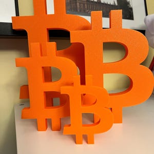 May include: Four orange Bitcoin symbols of different sizes are displayed on a white surface. The symbols are three-dimensional and stand upright. A framed picture of a building is in the background.